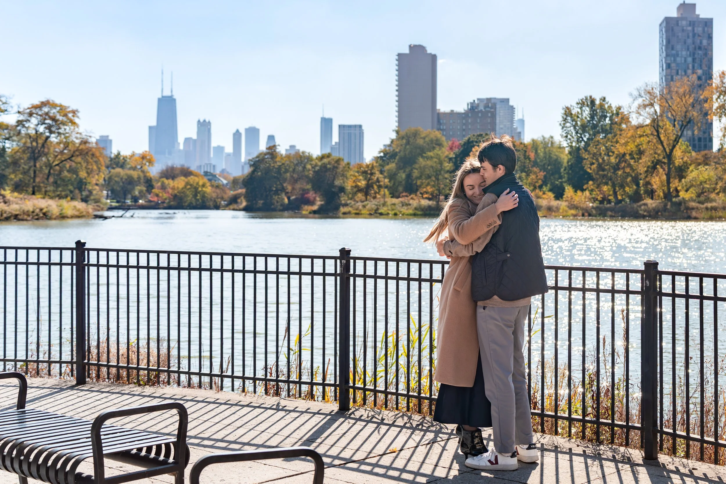 A couple hugging by a lake in a city park with skyscrapers in the background on a sunny autumn day.