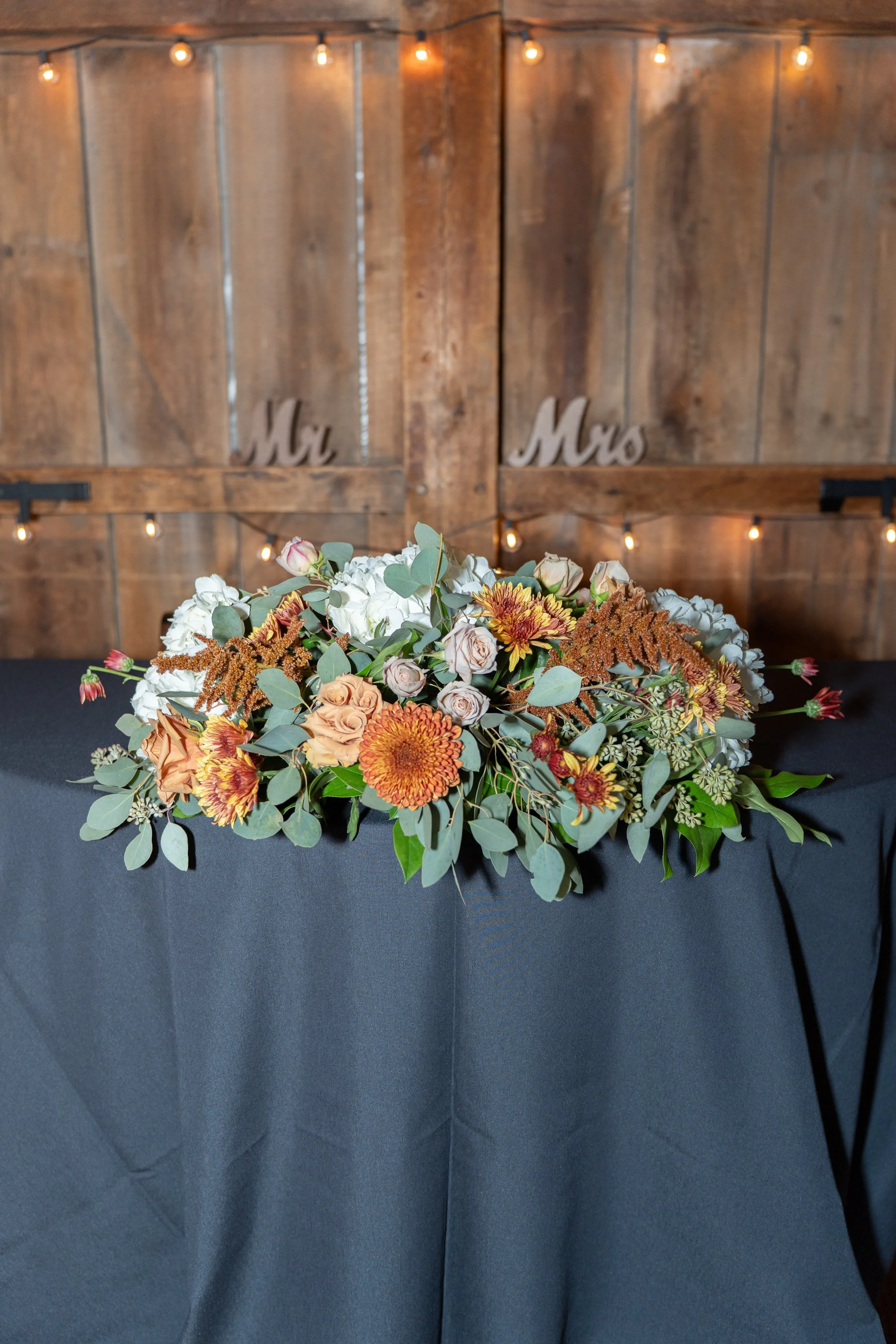A floral arrangement on a black tablecloth with a rustic wooden background and string lights, featuring signs reading 'Mr.' and 'Mrs.' in the background.