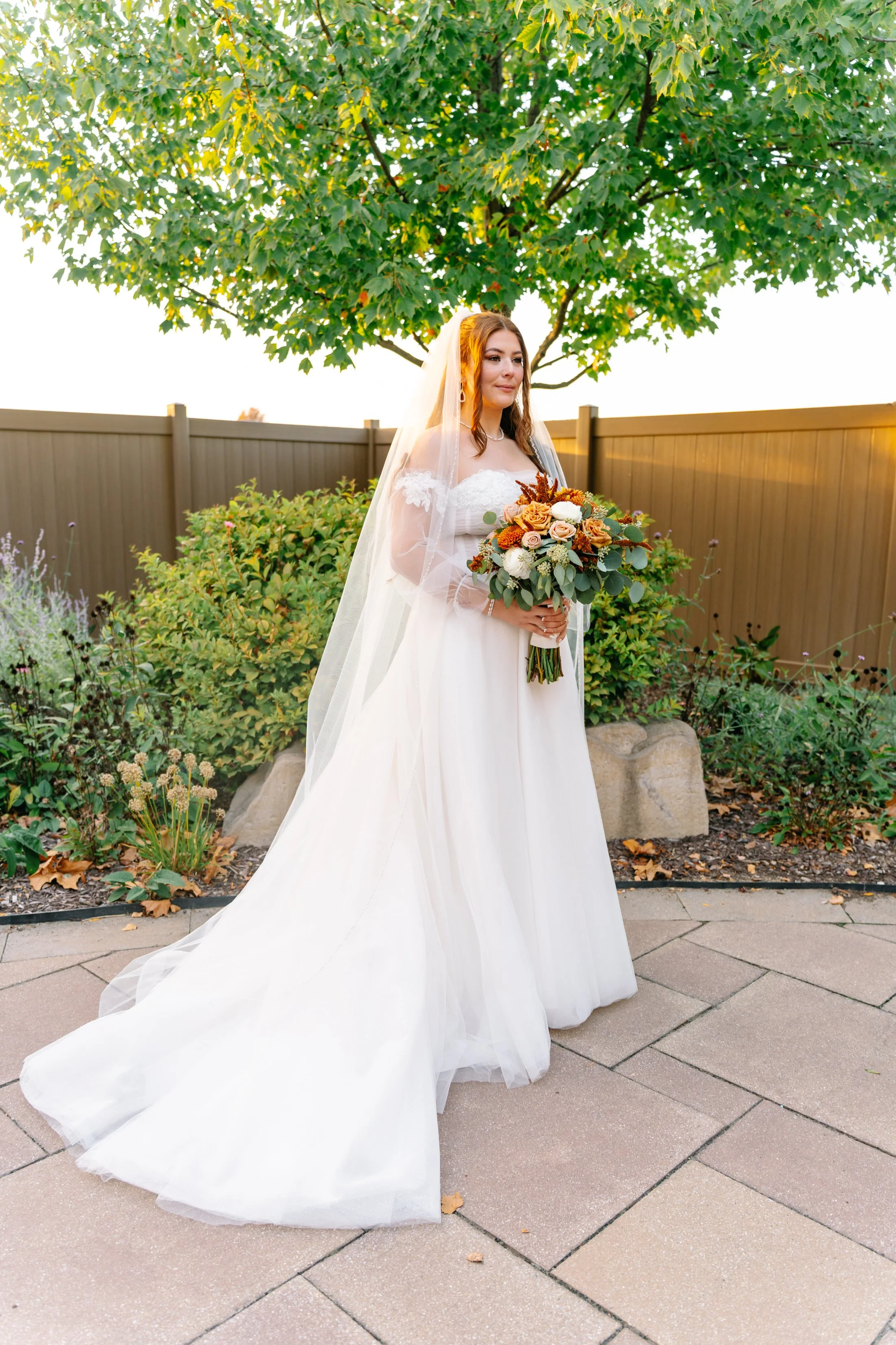 Bride in white wedding dress holding a bouquet of flowers outdoors, standing on a paved patio with greenery and a fence in the background.