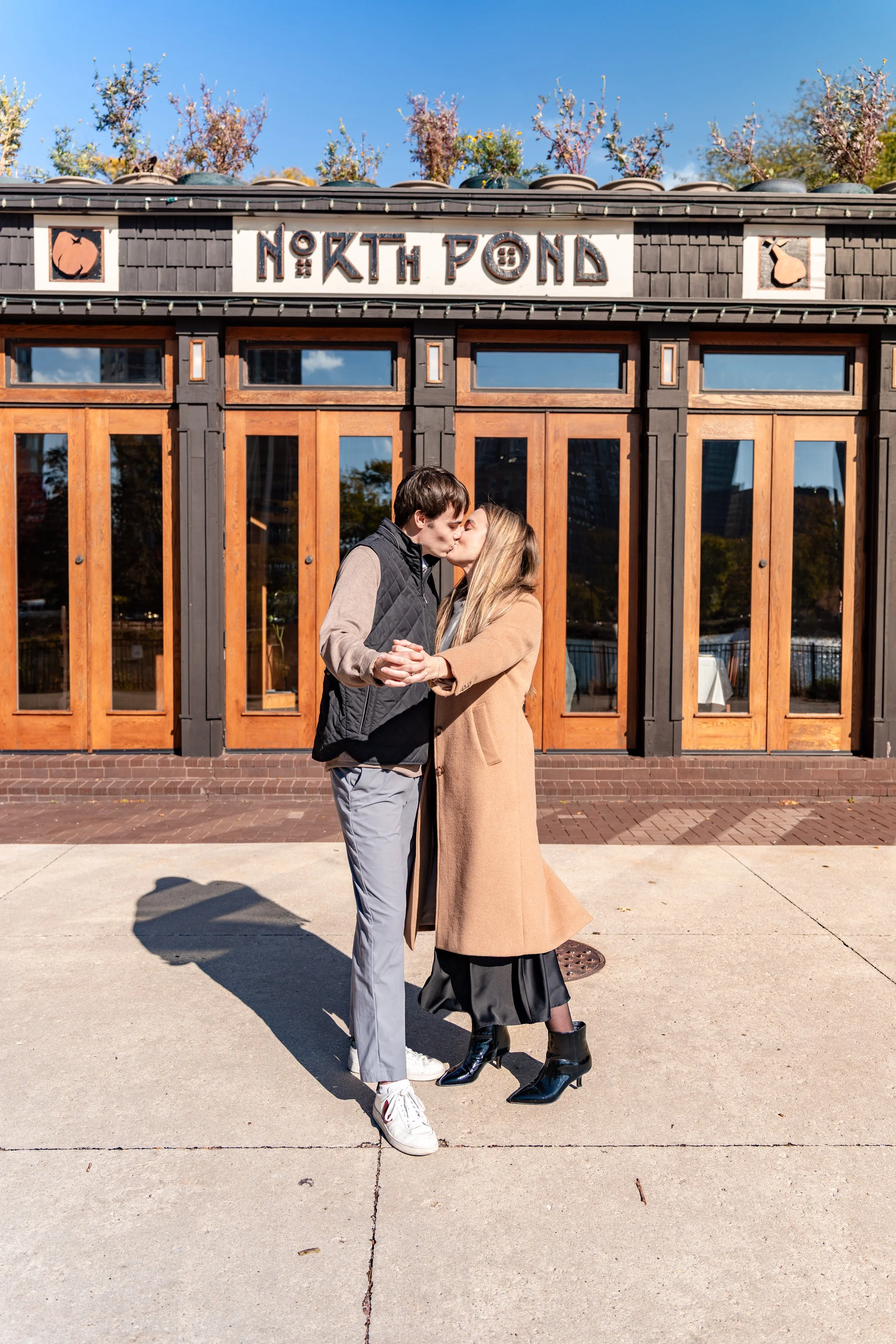 A couple kissing in front of a wooden building with a sign that reads 'North Pond' and decorative images of a pumpkin and a duck. The couple is holding hands and dressed in fall clothing.