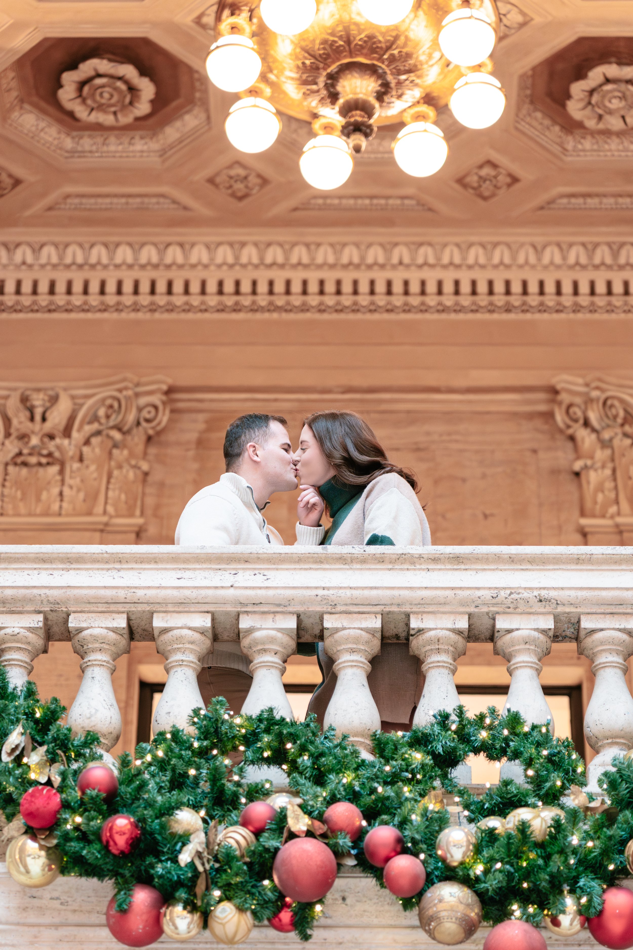 Couple kissing on a decorated balcony with Christmas garland and ornaments, inside a grand wooden hall with ornate ceiling and chandelier.