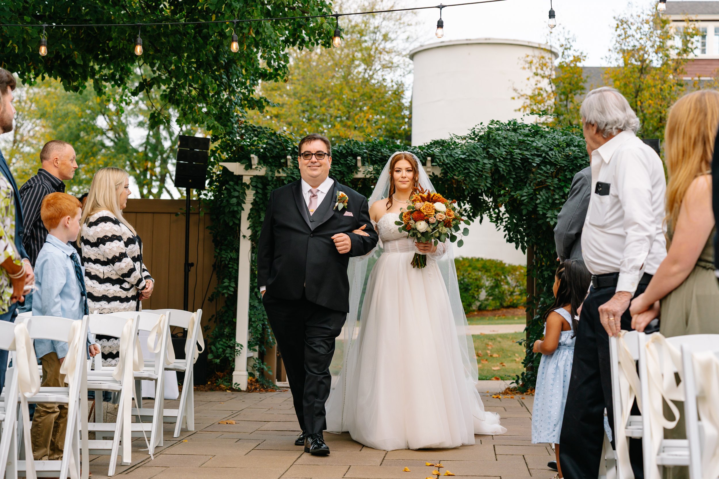 Bride walking down the aisle with her father during a wedding ceremony outdoors, with guests seated on white chairs on either side and string lights hanging above, greenery, and a white building in the background.
