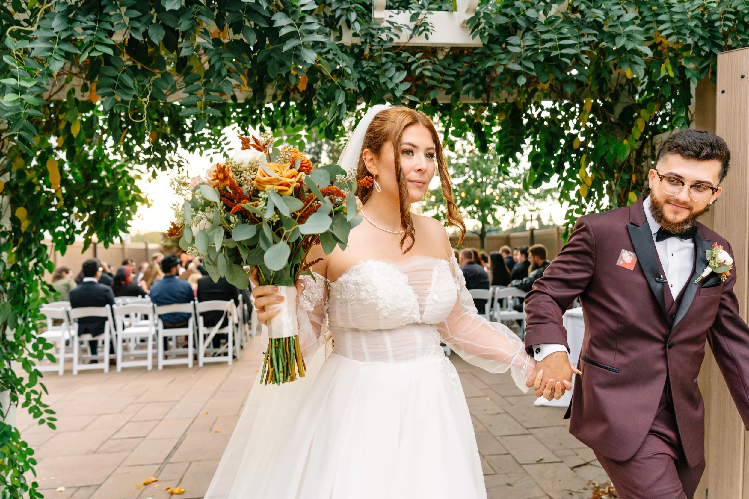 A bride and groom holding hands at their outdoor wedding ceremony. The bride is wearing a white wedding dress and holding a large bouquet of orange and peach flowers with greenery. The groom is dressed in a burgundy tuxedo with a black bow tie and gl