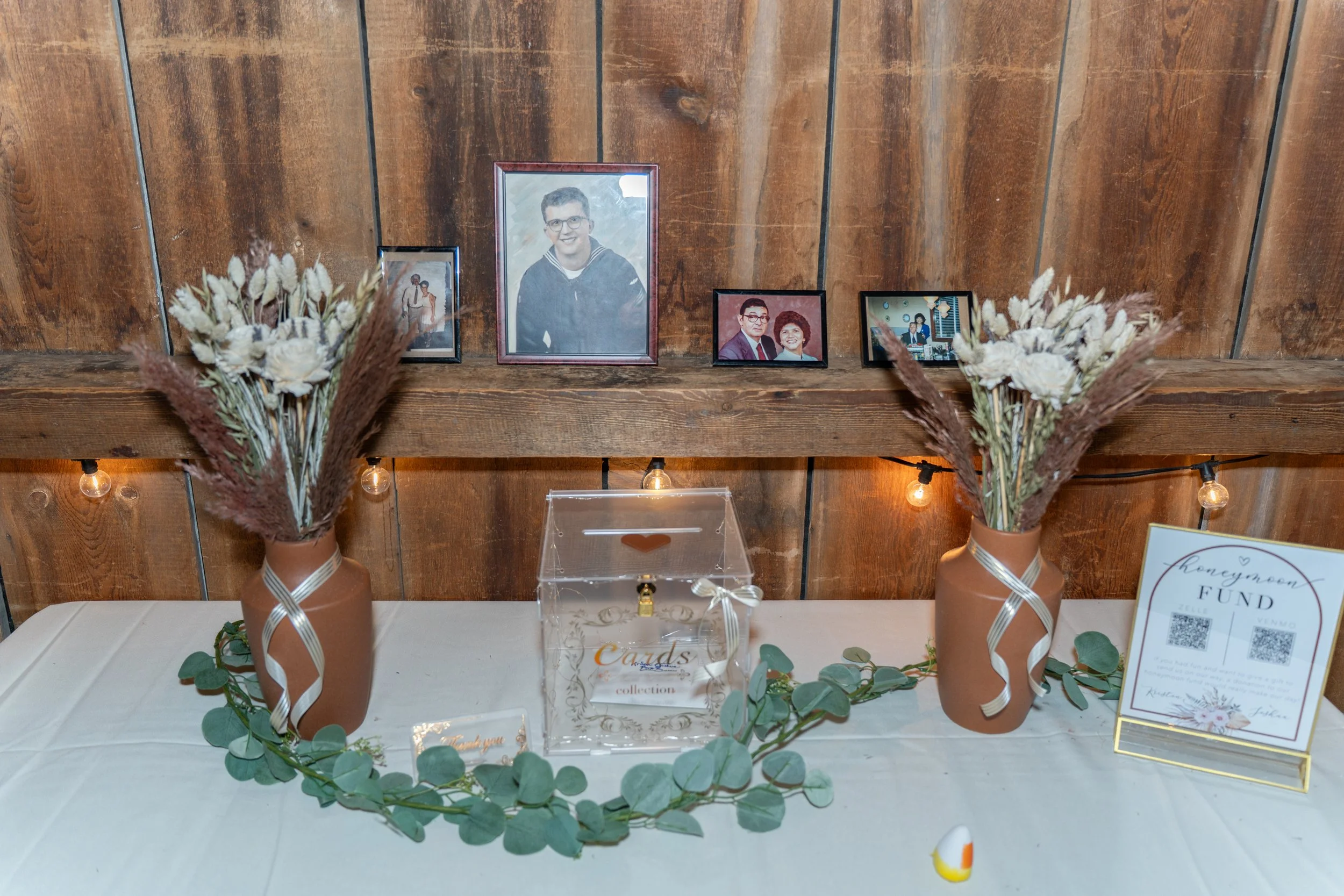 A wedding or memorial table decorated with two brown vases holding dried floral arrangements, a box labeled for cards, and a framed sign with a QR code. Behind the table, a wooden wall displays four framed photographs of different people.