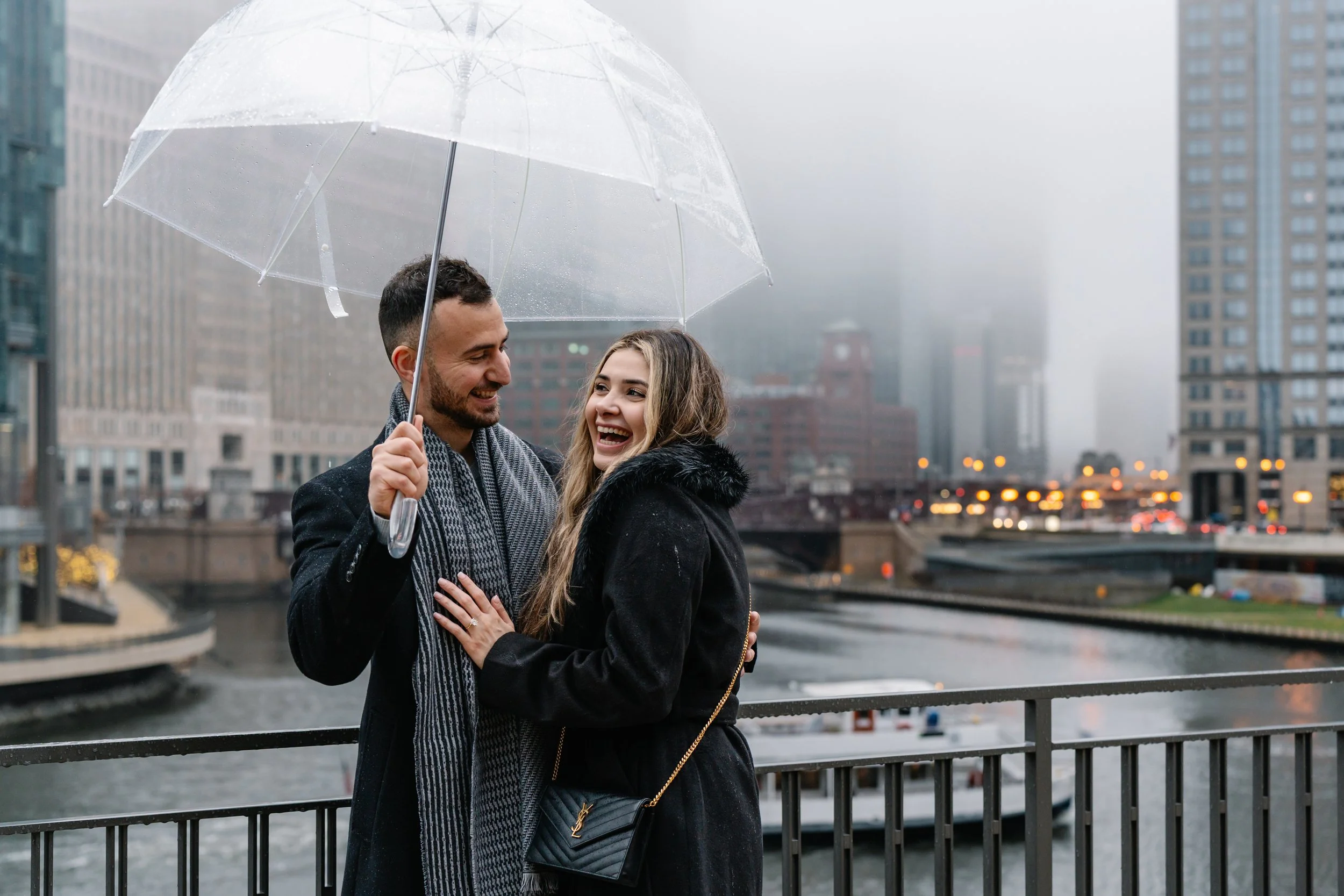 A couple sharing a laugh under a transparent umbrella on a rainy day in an urban area with tall buildings and a river in the background.