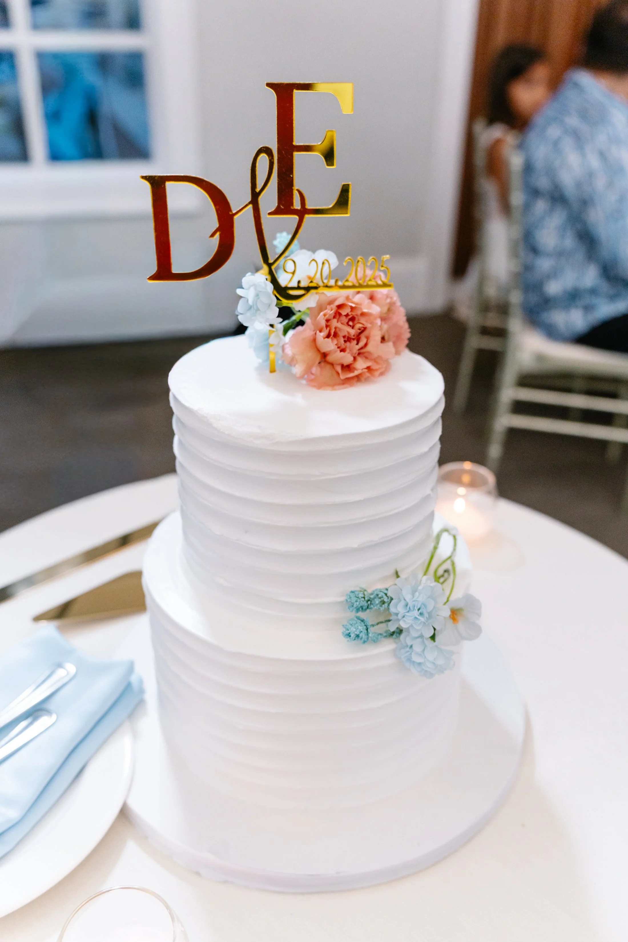 White, tiered wedding cake decorated with pink, white, and blue flowers, topped with gold cake topper featuring initials 'D' and 'E' and the date September 20, 2025.