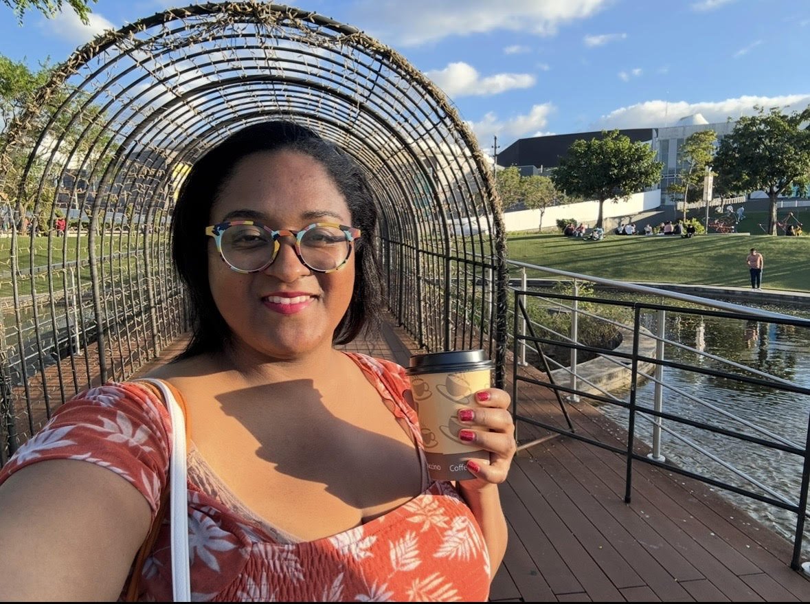 A woman taking a selfie outdoors near a pond with a bridge structure behind her, holding a coffee cup, dressed in a floral pattern red and white top, wearing round glasses, with a smile and sunglasses on her head.