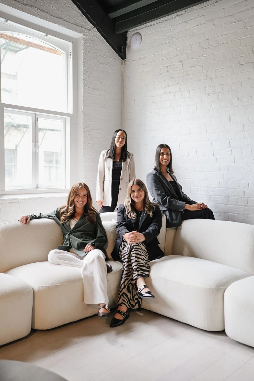 Four women in business attire sitting and standing on a cream-colored sofa in a minimalistic white brick room with large windows.