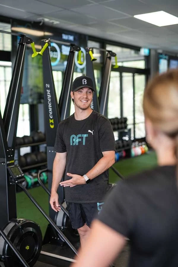 A fitness trainer talking to a woman in a gym with exercise equipment in the background.