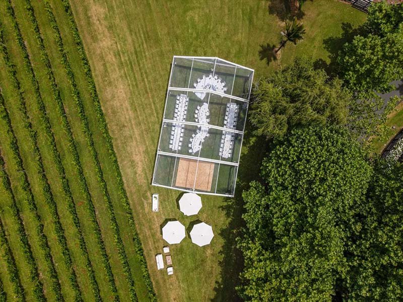 An outdoor event space with a glass tent set up on lush green grass next to a vineyard or farm field and tall trees, with white umbrellas and outdoor furniture nearby.