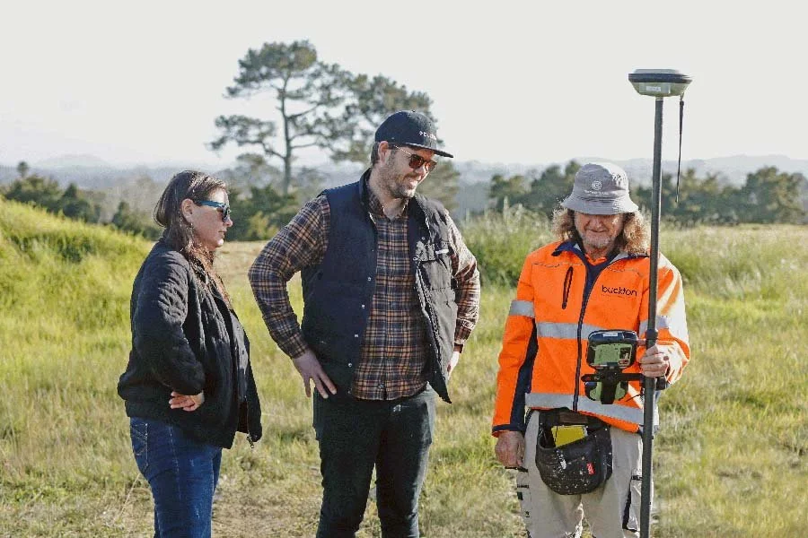 Three people outdoors in a grassy field, with one holding a surveying instrument and the others observing.