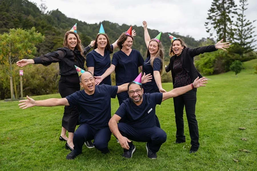 Group of seven people celebrating outdoors with colorful party hats, smiling and posing happily on a grassy field with trees and hills in the background.