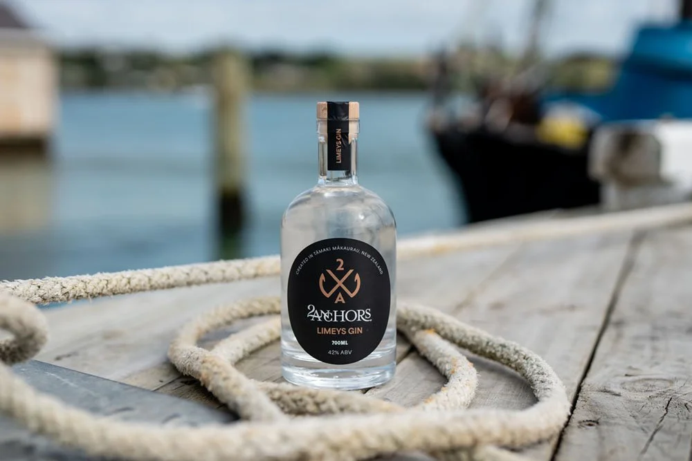 A bottle of Anchors Limeys Gin placed on a wooden dock near water, with boats and a bridge in the background.