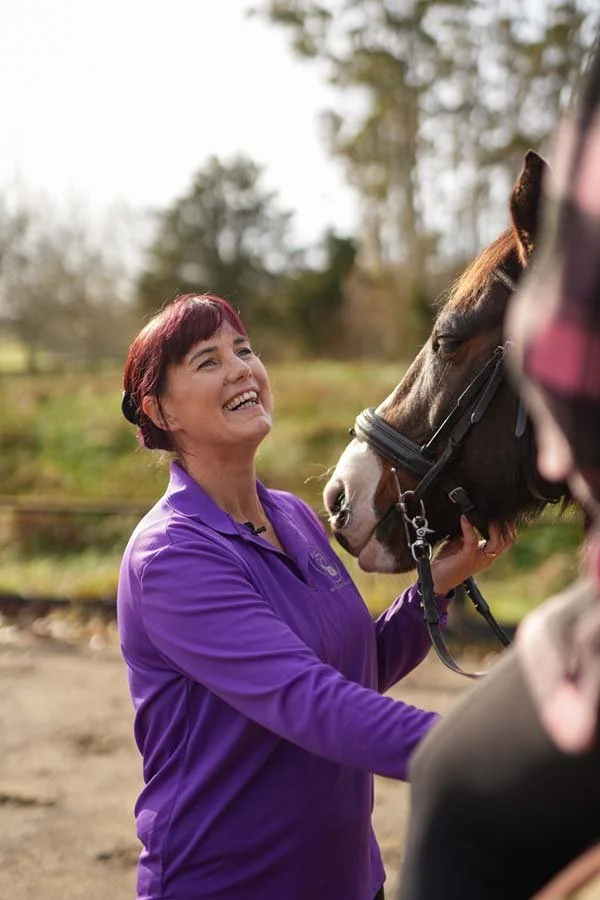 A woman with short red hair smiling at a horse during daytime outdoors.