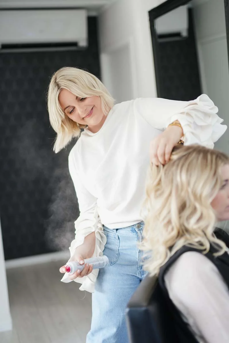 A hairstylist spraying hair with a spray bottle while working on a client's blonde hair in a salon.