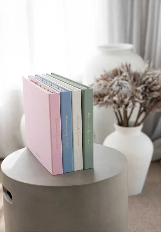 Four pastel-colored hardcover books standing upright on a round, light gray tabletop, with a white vase of dried flowers in the background near a window with sheer curtains.