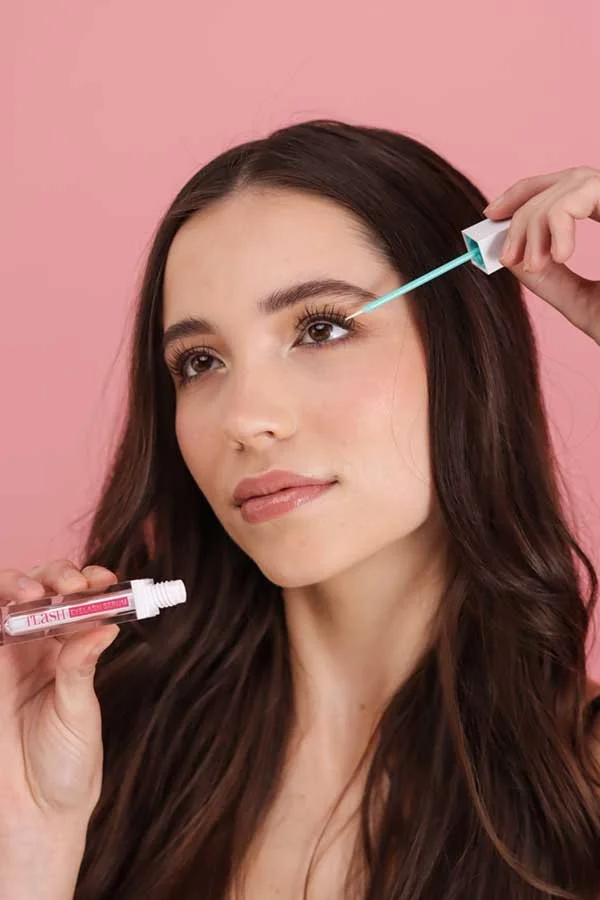 A woman applying eye cream with a roller applicator while holding a tube of eye treatment, with a pink background.