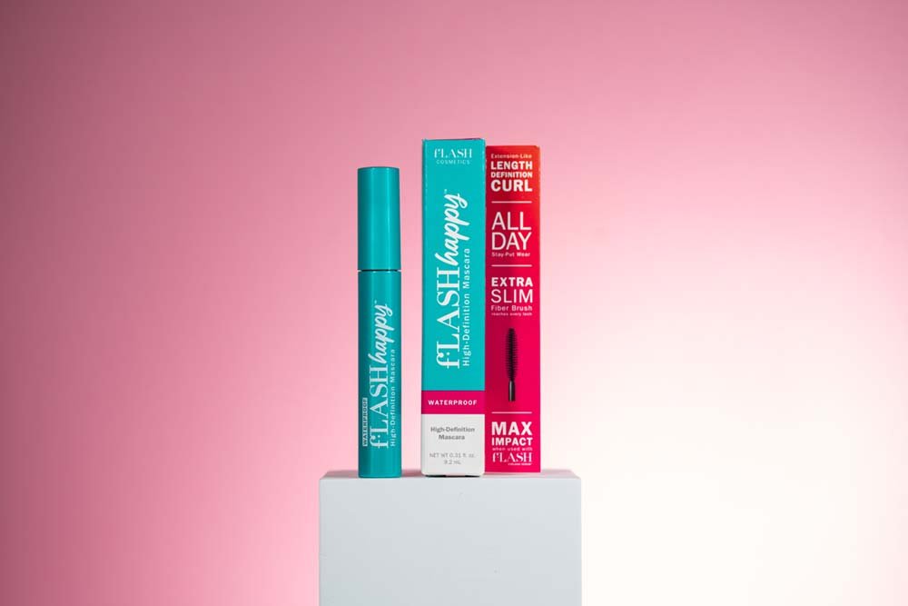 Blue tube of waterproof mascara, teal boxed mascara, and red and black box of fiber brush mascara displayed on a white pedestal against a pink background.