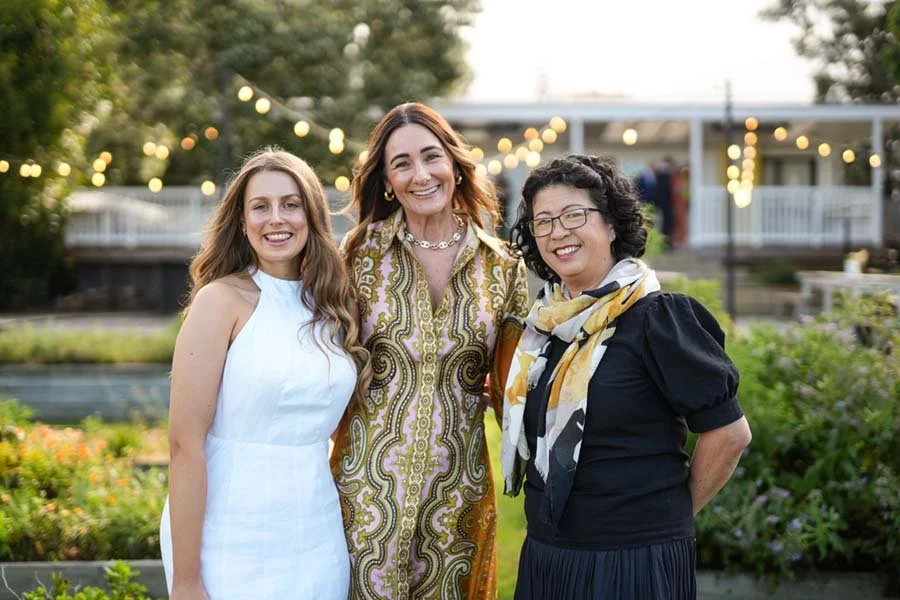 Three women standing outdoors at sunset, smiling, with string lights and greenery in the background.