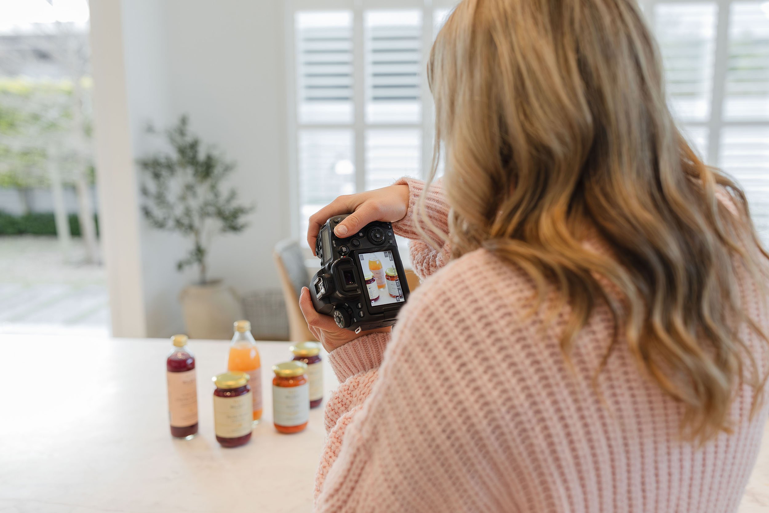 Tayla Keon taking a photo with a camera of bottles and jars of vitamins or supplements on a white table in a bright room with large windows and a potted plant in the background.