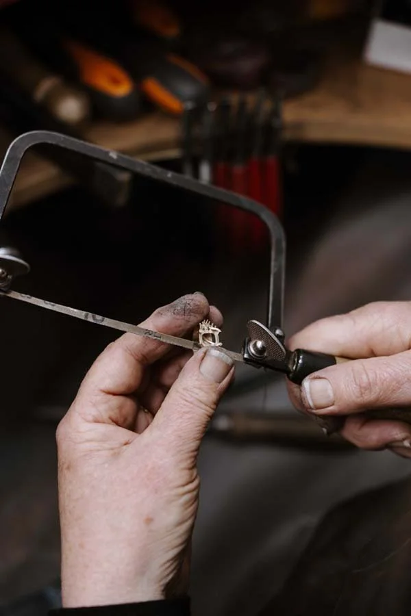 Person using a jeweler's saw to cut a small metallic object, possibly a ring, in a workshop setting.