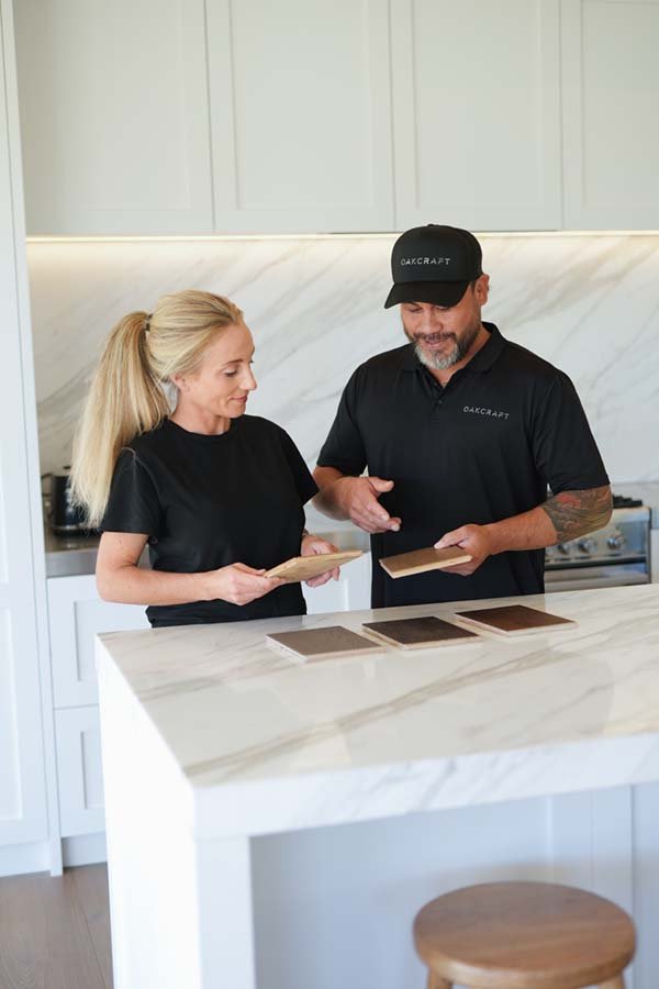 A man and woman in black shirts, with the man wearing a black cap, standing at a kitchen island examining color samples, with white cabinets and marble backsplash in the background.