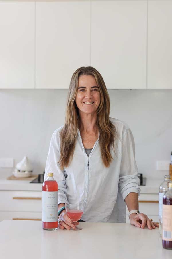 A woman with long brown hair smiling in a bright kitchen, holding a pink cocktail in one hand, with bottles on the counter.