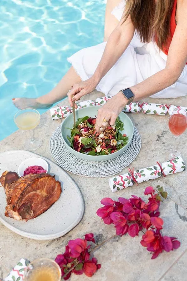 A woman sitting by a poolside with a meal of salad, grilled meat, and drinks, decorated with pink flowers and holiday-themed napkins.