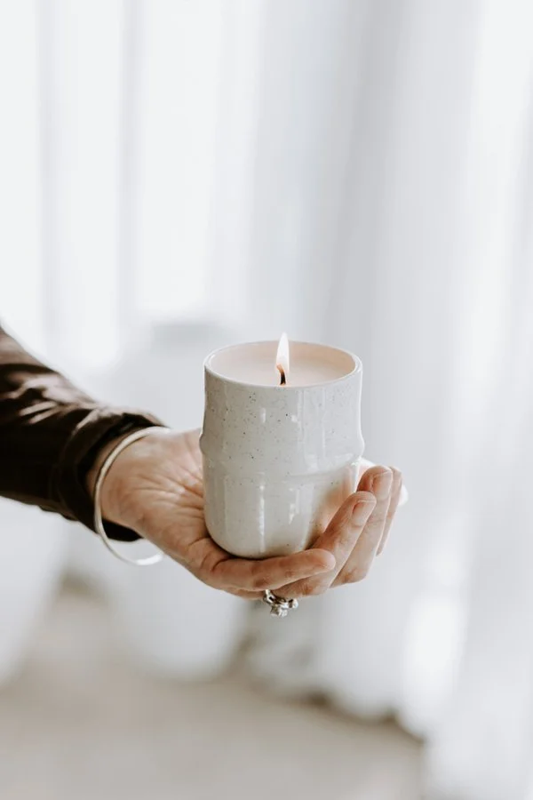 Person holding a white, ceramic candle holder with a lit candle inside, in front of a bright window with sheer curtains.