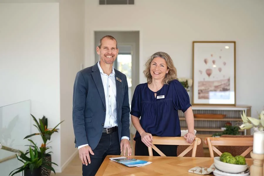 A man in a suit and a woman in a blouse stand in a bright, modern room, smiling at the camera. They are inside a home or office, with a wooden table, chairs, and a framed picture on the wall behind them.