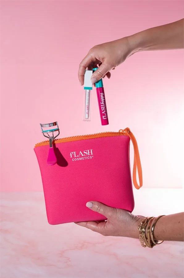 Hand holding a pink cosmetic pouch with orange zipper, labeled 'fLASH COSMETICS'. Another hand holds a pink and blue mascara tube and a clear lip gloss tube with blue cap above the pouch. A pink and blue eyelash curler is clipped onto the pouch.