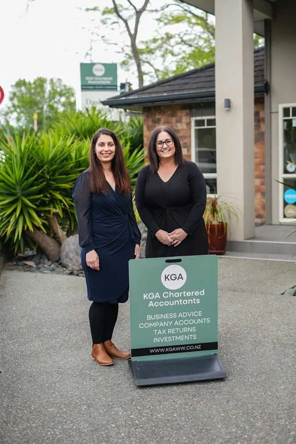 Two women standing outdoors next to a sign for KGA Chartered Accountants, with business services listed. One woman wears a navy dress, the other a black dress, both smiling.