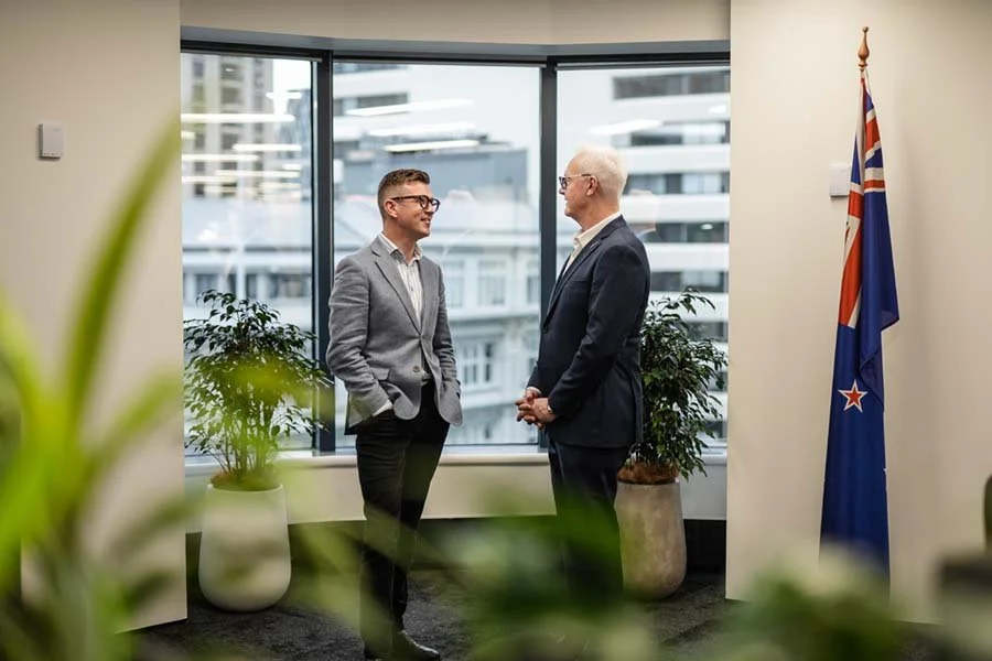 Two men in suits having a conversation in an office with large windows and plants, one young with glasses and the other older with white hair, next to a New Zealand flag.