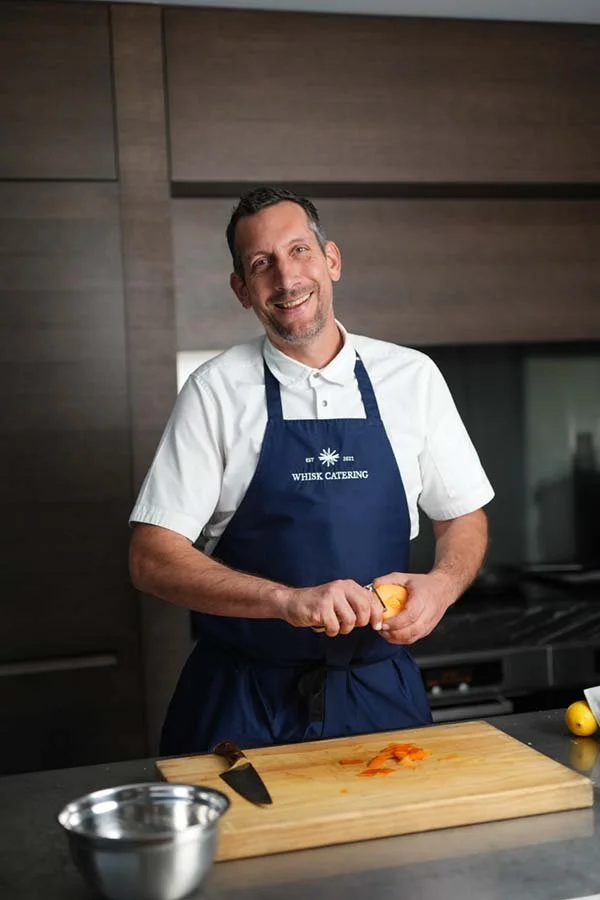 Man in a white shirt and blue apron chopping an orange on a wooden cutting board in a modern kitchen.