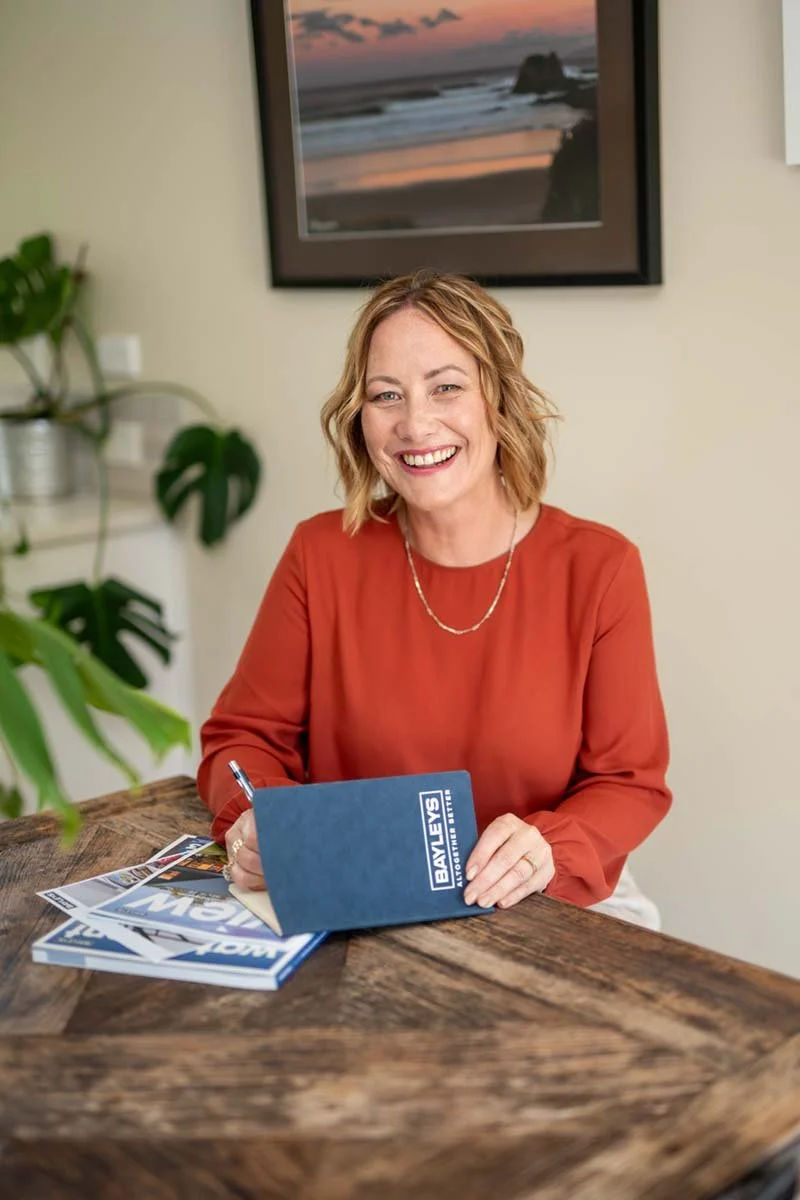 A woman with shoulder-length blonde hair sitting at a rustic wooden table, smiling and holding a blue folder with "Bayleys" written on it, with some magazines in front of her. A framed sunset photo and green plants are in the background.