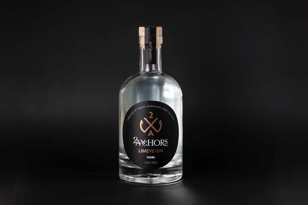 A clear bottle of 2 Anchors Limeys Gin with a black and copper label, standing against a black background.