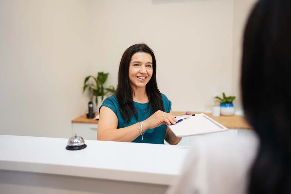 A woman at a reception desk smiling and holding a clipboard, with another person partially visible in the foreground.