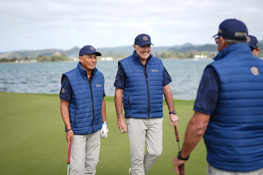 Four men in blue vests and caps standing on a golf course talking and smiling near water.