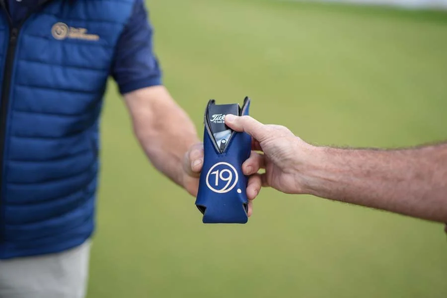 A person in a blue vest is handing a golf scorecard holder with the number 19 and a golf scorecard inside to another person on a golf course.