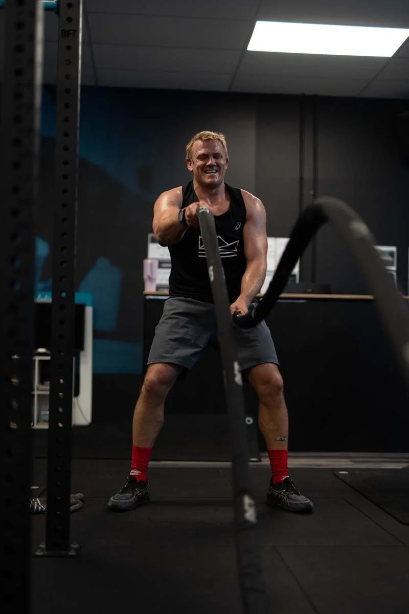 A man exercising with battle ropes in a gym, smiling and wearing workout clothes, including a black tank top, gray shorts, red socks, and running shoes.