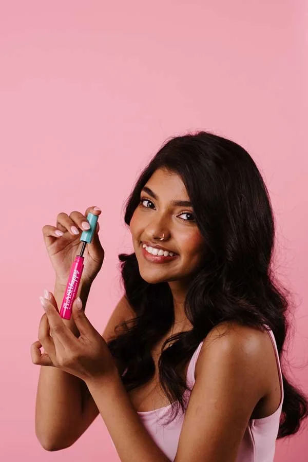 A young woman with long dark hair, smiling, holding a pink and teal vape pen against a pink background.