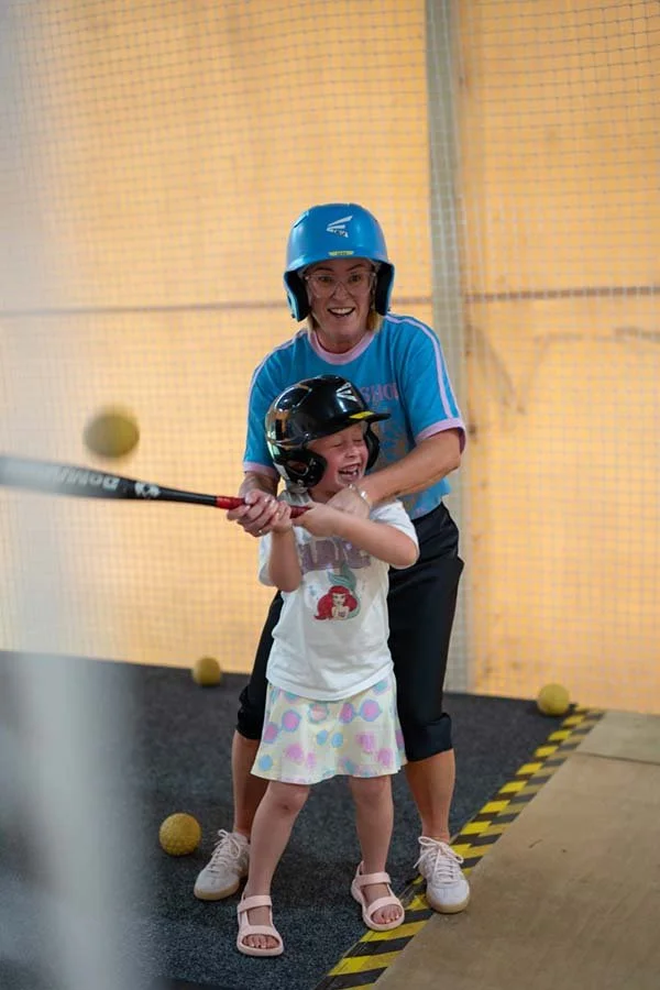 A young girl and an adult woman are playing indoor baseball. The girl is wearing a helmet and holding a bat, and the woman is standing behind her, helping her swing. The girl is laughing and smiling, and they are surrounded by yellow balls on the bla