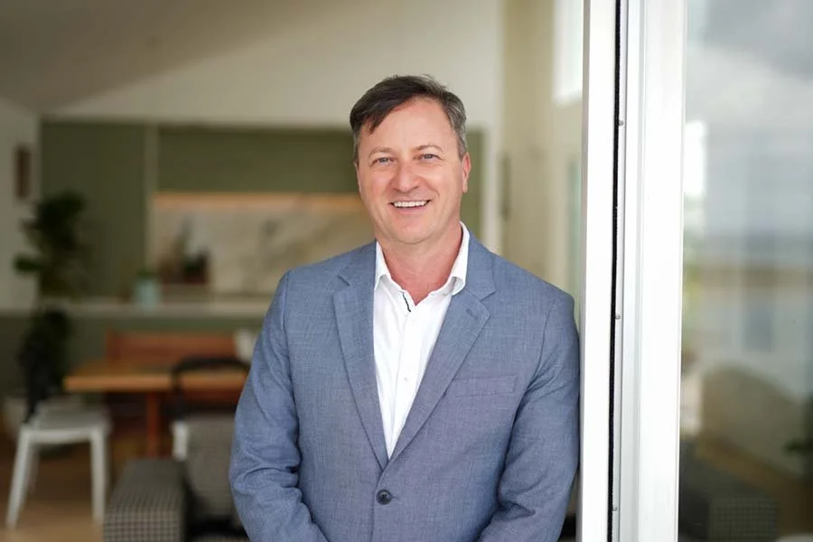 A smiling man in a gray suit and white shirt standing near a sliding glass door in a modern apartment.