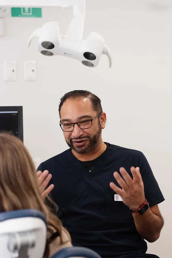 A male dentist or dental professional wearing glasses and black scrubs talking to a patient in a dental office, with dental light above.