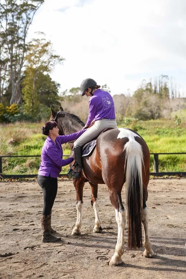 A woman in purple helping a woman in purple ride a brown and white horse outdoors on a sunny day.