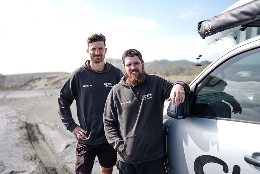 Two men standing outdoors near a vehicle on a dirt road with open landscape in the background. One man leans on the vehicle's door and the other stands with hands on hips. Both are wearing hoodies.