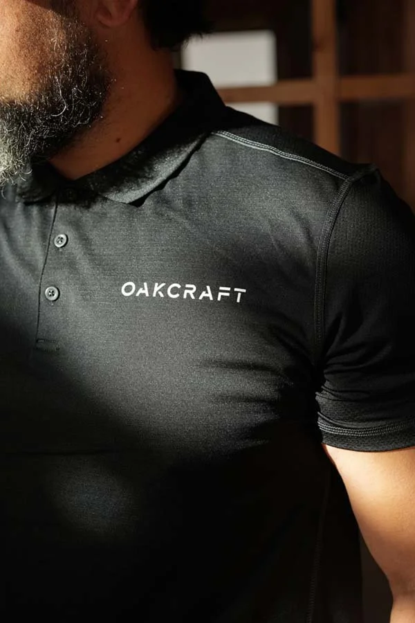 Close-up of a person wearing a black OAKCRAFT sports shirt with a partial view of their face, beard, and shoulder, in an indoor setting.
