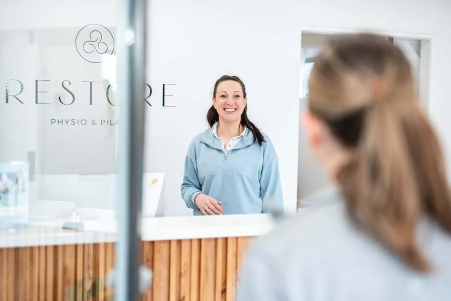 A woman in a light blue jacket smiling at a front desk in a health or wellness facility.