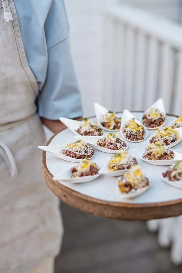 Person holding a round tray of small appetizers served in white spoons, garnished with yellow and white toppings.