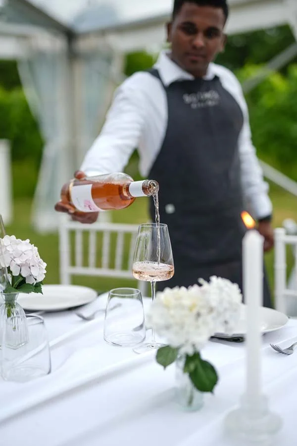 A waiter pouring rosé wine into a glass on a decorated outdoor dining table with white flowers and candles.